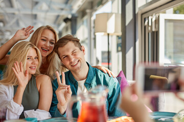 portrait of three young best friends sitting in cafe, taking photo, woman hold mobile phone in hands and take photo of friends, smiling