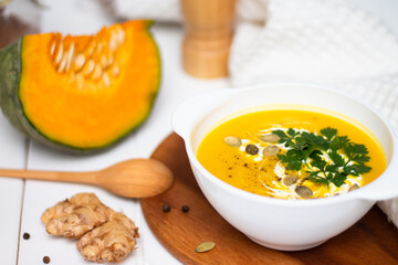 A plate of yellow pumpkin cream soup with cream, herbs and seeds on a white table near raw pumpkin and a wooden spoon.