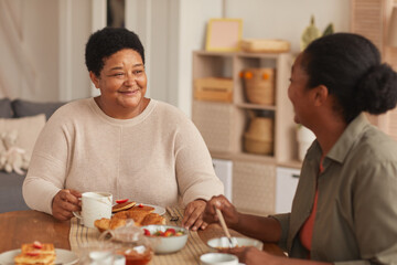 Warm toned portrait of happy African-American woman sitting at dining table with daughter while...