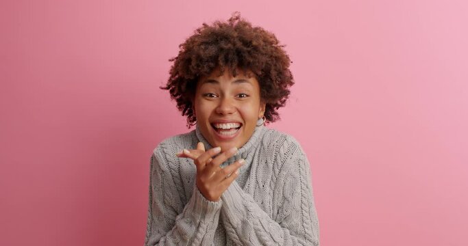 Happy dark skinned young woman makes triumph gesture says yes I did it makes victory dance glad to achieve great results dressed in knitted sweater poses against rosy background. Success concept