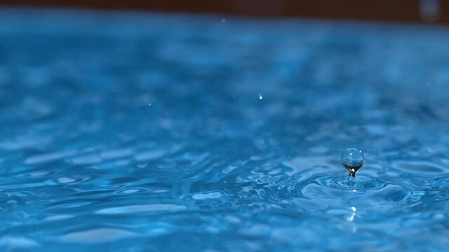 SLOW MOTION, MACRO, DOF: Glassy drops of rain begin to fall into a pool at the beginning of a torrential rainstorm. Crystal clear raindrops create a ripple effect on the surface of an empty pool.