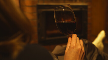 Woman with glass of red wine is resting by the fireplace.