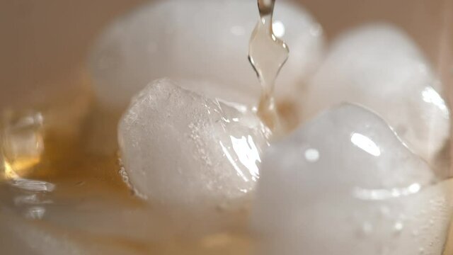 SLOW MOTION, MACRO, DOF: Tasty bourbon is poured over the shiny melting round pieces of ice sitting in a crystal glass on the counter of a restaurant. Decadent whiskey trickles over the ice cubes.