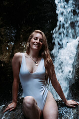 Vertical shot of a sexy woman in a wet one-piece swimsuit posing on a rock with a waterfall behind