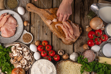 Chef hands cuts with knife chicken fillet on wooden chopped board with variety of ingredients background. Concept of cooking process. Backstage of preparing tasty meal. View from above. Flat lay.