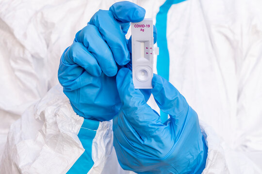 A healthcare worker covered by a white coverall and with blue gloves shows a rapid test for the control of a possible contagion from covid-19 - Powered by Adobe