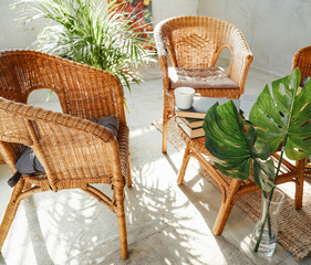 Warm and classy hotel room there are bamboo chairs with pillows and table with coffee cup and books around plant in sunshine.