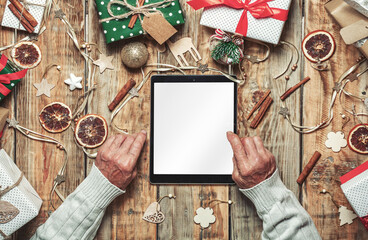 Hands of elderly senior with tablet, blank empty screen,
on table with christmas gifts and decorations