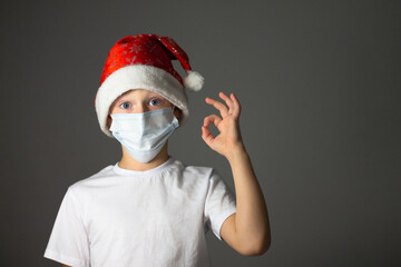 7-10 boy in a Christmas cap and medical mask shows OK. Christmas and new year during coronavirus pandemic concept.