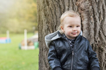 Adorable little boy dressed  in a leather jacket is sticking out his tongue outdoors.