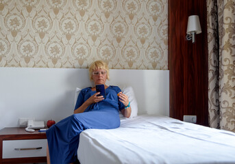 Relaxed woman sitting on the bed in the hotel, holding a smartphone or shop online to check mobile application or social network. Selective focus. - Powered by Adobe