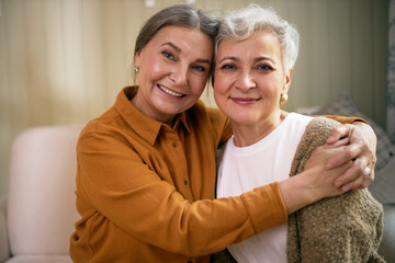 Portrait of two cheerful senior Caucasian female friends embracing, looking at camera with happy smile, glad to see each other after long time. Joyful elderly sisters posing indoors, hugging
