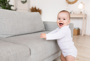 Fototapeta premium Cute Little Baby Girl Smiling Standing Near Sofa At Home