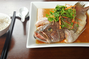 Steamed whole Grouper Fish in Soy Sauce of Chinese cooking style and garnish with shredded ginger and parsley, served on wooden table.