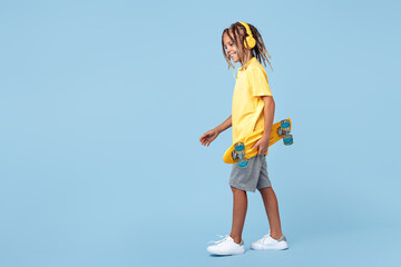 Modern little boy with african dreads with headphone and skateboard posing over blue background.
