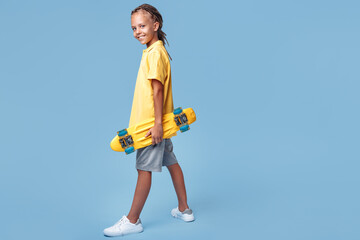 Fototapeta na wymiar Full lengh image of stylish little boy with african dreads with headphone and skateboard posing over blue background.
