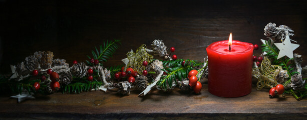 Red candle and Christmas decoration with rose hips, branches and cones on a dark rustic wooden...