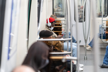 Afro-American passenger man sitting in subway train, wear face medical mask to protect yourself from contact with flu virus, covid-19, using mobile phone, listens to music with wireless headphones. 