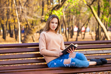 Beautiful blonde girl with a book sits on a bench in an autumn park