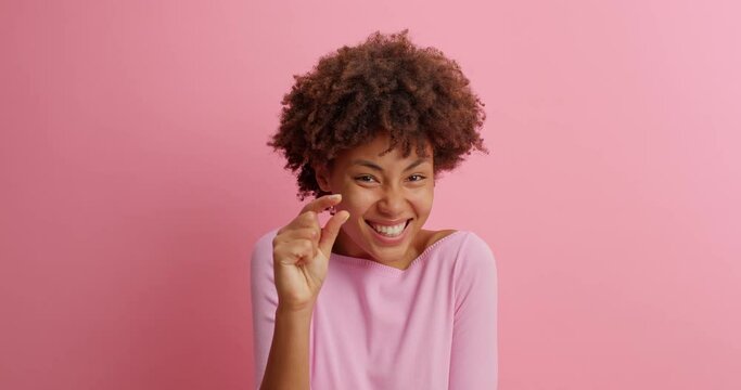 Young brunette young woman with curly Afro hair shows very tiny object shapes small item needs some more dressed in casual clothing poses against pink background. Minimum size. I need not much