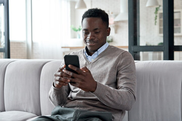 Smiling african american man holding smart phone using social media apps sitting on couch at home....