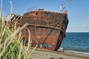 old rusty and rotten ship wreck with holes in its carcass, lying behind long gras on the beach of...