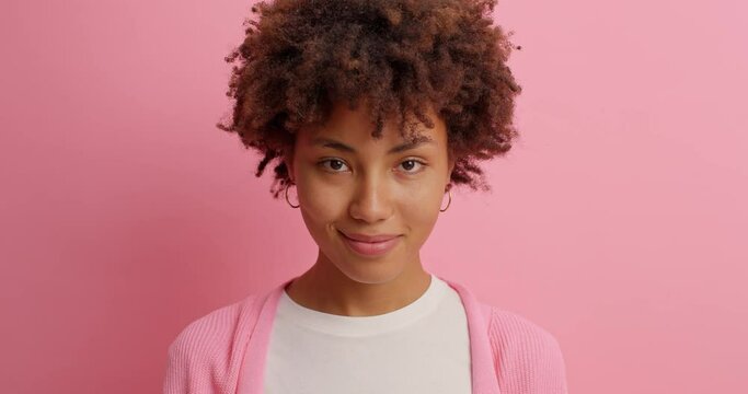 Beautiful calm young woman has Afro hair looks seriously then smiles gently gazes directly at camera has pleasant talk with interlocutor poses against pink background. Face expressions concept