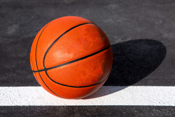 orange striped basketball ball stands on the white line marking the playing field with asphalt tarmac, sports object lit by sun light on playground, nobody.