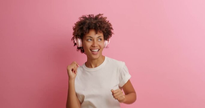 Merry curly haired woman has upbeat mood dances carefree listens pleasant music in headphones moves with rhythm sings song keeps hand near mouth as microphone wears basic white t shirt poses indoor
