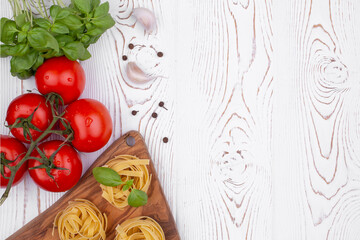 Top view raw tagliatelle pasta with fresh basil, garlic and tomatoes on a rustic white table, flat lay, copy space.