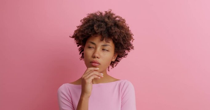 Thoughtful Afro American woman leans on hand has bored pensive expression tries to gather ideas for future stands unaware indoor dressed casually models against pink background. Slow motion.