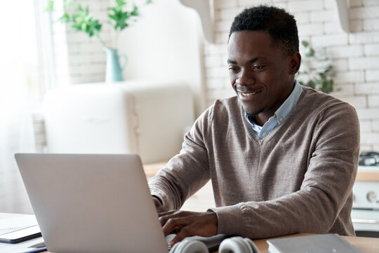 Smiling professional black business man using laptop computer working from home office. Happy african male customer makes ecommerce delivery order from supermarket typing on pc sits at kitchen table.