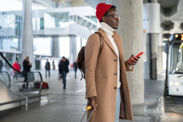 Trendy Afro-American traveler millennial man in beige coat, red hat with yellow suitcase holding...