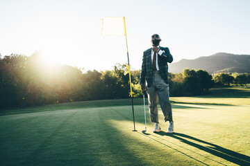 A stately fashionable bearded black guy in a costume and sunglasses is straightening his tie while standing on a golf field with a club in hand on a warm sunny evening near the hole with a yellow flag