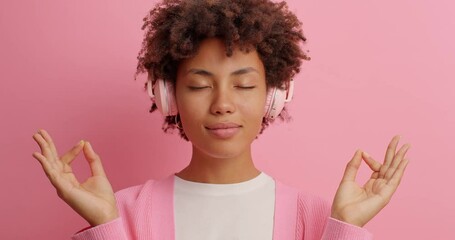 Relaxed beautiful Afro American woman listens music in stereo headphones meditates and tries to relax enjoys calm pleasant melody closes eyes poses against rosy background. Peaceful atmosphere