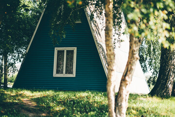 View of a fragment of a summer cottage with a triangle roof to the ground, in the countryside and a birch grove, lit by the summer sun, with a green lawn and two twisted tree trunks in the foreground