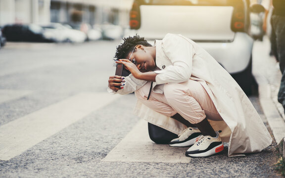 A young fancy black female tourist in a white stylish trench is squatting on a pedestrian crossway, near the road while trying to take a pic on a camera of her smartphone from the bottom