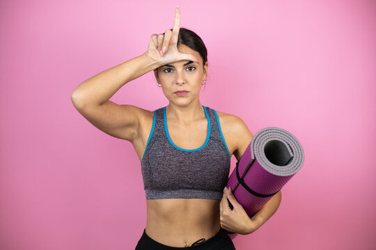 Young beautiful woman wearing sportswear over isolated pink background holding a splinter in her hand making fun of people with fingers on forehead doing loser gesture mocking and insulting.