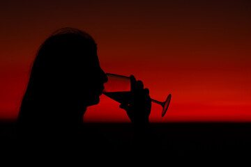 the woman holding the glass of wine in her hand and put it to her mouth. drinking wine with a sunset, silhouette of a woman drinking wine at sunset.