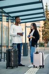 A joyful multiracial couple checks their boarding passes and departure time at a bus stop near the airport.