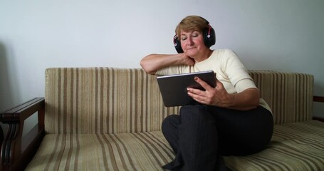 Senior woman connected with tablet and headphones, relaxing in sofa - Powered by Adobe