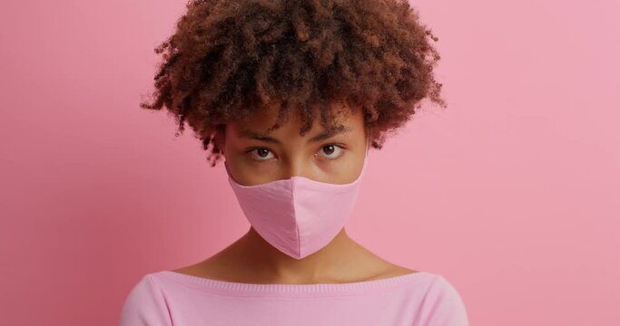 Serious African American woman concentrated down wears protective mask against covid virus has calm expression prevents disease spread isolated over pink background. Coronavirus and self isolation