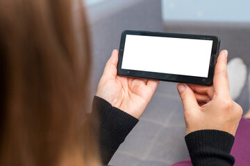 A female is holding and using smartphone with white blank screen. Woman lies on sofa and holds phone with white blank screen