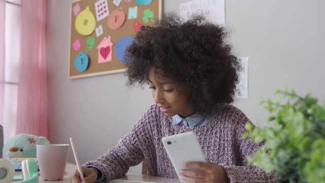 African american child kid school girl holding phone distance learning class using mobile application, watching online tutoring lesson, video calling in app making notes studying at home in classroom.