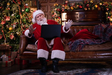 Obraz premium Kids and Santa in medical masks. Girl and Santa Claus reading Christmas book sitting near Christmas tree at home. Happy New Year Covid 2021.