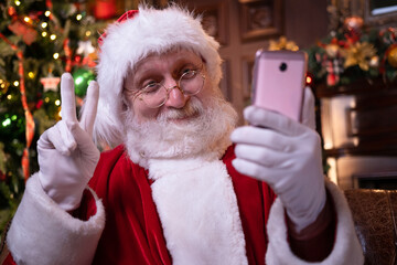 Christmas online congratulations from Santa. Santa Claus using mobile phone for distance Christmas call people and kids. Sitting near Christmas tree at home. Happy New Year Covid 2021.