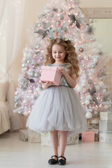 little girl in grey dress, hoop with deer antlers, with pink gift box in hands against the background of white Christmas tree. child with present for the new year. happy childhood. vertical