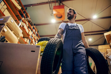 Low angle view of tattooed bearded hardworking smiling employee relocating tires while walking in...