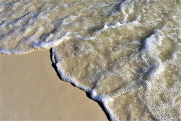 A wave rolls back from the shore, exposing light, clean sand and foaming at its edge, viewed from above