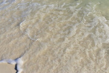 A wave rolls back from the shore, exposing light, clean sand and foaming at its edge, viewed from above